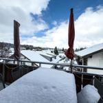 Schneebedeckter Balkon mit Tisch und Stühlen, Blick auf verschneite Dächer und Berge.