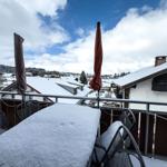 Schneebedeckter Balkon mit Tisch und Stühlen, Blick auf schneebedeckte Dächer und Berge.