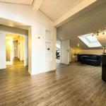 Wohnung mit Holzboden, Dachschrägen und Fenstern. Ein dunkler Sofa und eine Küche sind sichtbar.