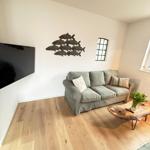 Living room with sofa, TV, and wooden table. Wall decoration and windows visible.