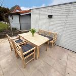 Terrace with wooden table, four chairs, and bench. Background: house wall and garden.