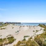 Strand mit Sanddünen, Häusern und Blick aufs Meer unter blauem Himmel.