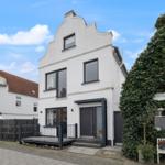 Weiße Hausfassade mit dunklen Fenstern und Eingang. Rechts wächst Efeu an einem Gebäude.