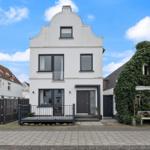 Weißes Haus mit dunklen Fenstern und Eingangstür. Vordere Terrasse und gepflasterter Hof.