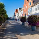 Straße mit Blumenkübeln und Caféterrassen unter klarem Himmel.