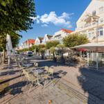 Außenbereich mit Tischen, Stühlen und Sonnenschirmen entlang einer Straße mit Bäumen und Gebäuden.