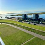 Hafenansicht mit modernen Gebäuden und Parkplatz. Grünflächen und Wasser im Hintergrund.