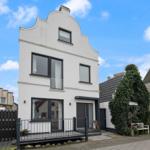 Weiße Hausfassade mit schwarzem Fensterrahmen und Vordereingang. Gartenbereich mit Bänken und Pflanzen.