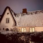Schneebedecktes Haus mit strohgedecktem Dach und beleuchteten Fenstern.