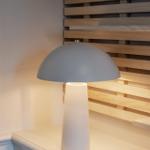 Eine Tischlampe mit grauem Schirm steht auf einer weißen Oberfläche vor einer Holzleistenwand.
