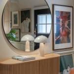 Rundes Spiegelbild, zwei weiße Lampen und ein Holzschrank mit Bildern an der Wand.