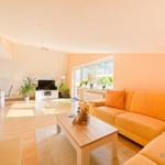 Wohnzimmer mit orangefarbenem Sofa, Holztisch und TV. Fenster mit Pflanzen. Holzboden und helle Wände.