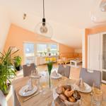 Esszimmer mit Tisch, Stühlen und Blumen. Im Hintergrund Wohnzimmer mit Sofa und Fenster.