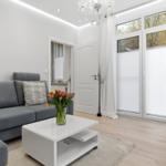 Wohnzimmer mit grauem Sofa, weißem Couchtisch und Blumen. Große Fenster mit weißen Vorhängen.