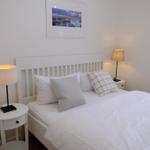 Schlafzimmer mit weißem Bett, zwei Nachttischen und Lampe. Wandbild zeigt Hafen.