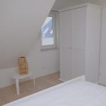 Schlafzimmer mit weißem Bett, Kleiderschrank und Fenster mit Blick auf Dach.