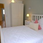 Schlafzimmer mit weißem Bett, Nachttisch und Kleiderschrank. Holzboden und Tür zum Flur.