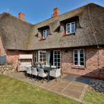 Haus mit Strohdach, Terrasse und Garten. Tisch mit Stühlen und Grill.
