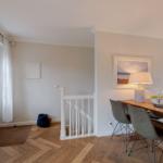 Dining area with wooden table, chairs, and lamp. Light walls, wooden floor, and staircase visible.