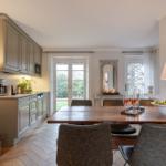 Dining room with wooden table, chairs, and kitchen area. Windows with garden view.