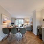 Dining area with table and chairs, living area with sofa and window, kitchen with countertop and coffee machine.