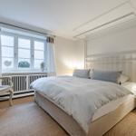 Bedroom with bed, nightstand, and chair. Window with curtains and radiator.