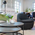 Living room with sofa, armchair, and dining table. Wooden floor and windows visible.