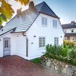 White villa with gray roof and paved driveway. Garden with stone wall and flowers.