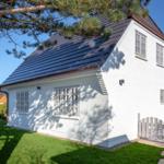 White villa with gray windows and dark roof. Green lawn and tree in front of the house.