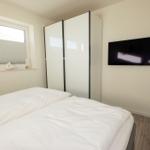 Schlafzimmer mit Doppelbett, Kleiderschrank und Wandfernseher. Fenster mit Jalousie.