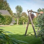 Hinter einem grünen Garten steht ein hölzerner Schaukelstuhl. Im Hintergrund ist ein Haus mit Dach zu sehen.