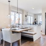 Dining area with table, chairs, and kitchen island. Wooden floor and large windows.
