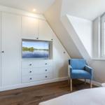 Bedroom with blue armchair, TV, and window. Wooden floor and white cabinets.