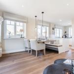 Dining area with table, benches, and windows. White wall and wooden floor.