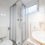 Badezimmer mit Dusche, Waschbecken und Toilette. Weiße Fliesen und Fenster.
