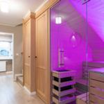 Sauna mit Glaswand und Holzbänken, beleuchtet mit lila Licht. Nebenraum mit Fenster und Holzboden.