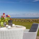 Rundtisch mit Blumen und Tassen im Freien, Blick auf Meer und Gras.