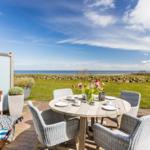 Terrasse mit Tisch, Stühlen und Meerblick. Strandkorb und Blumenarrangement sichtbar.