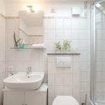 Badezimmer mit Dusche, Waschbecken und Toilette. Weiße Fliesen und Glasregal.