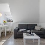 Wohnzimmer mit grauem Sofa, weißem Tisch und Stühlen, Flur mit Tisch und Blumen.