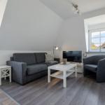 Wohnzimmer mit grauem Sofa, weißen Möbeln und Holzboden. Fenster bieten Blick auf Dachziegel.