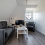 Wohnzimmer mit Sofa, Sessel, Couchtisch und Fernseher. Holzboden und weiß gestrichene Wände.