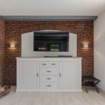 Wohnzimmer mit Kamin, Fernseher und weißen Schränken an Backsteinwand.