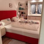 Schlafzimmer mit rotem Bett, Bücherregal und Fenster mit Vorhängen.
