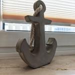 Ein grauer Anker mit Seil steht auf einer Steinplatte vor einem Fenster.