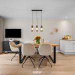 Esszimmer mit Holztisch, vier Stühlen und moderner Beleuchtung. Küche mit weißen Schränken im Hintergrund.