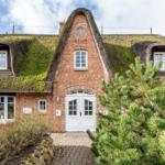 Haus mit strohgedecktem Dach, weiße Tür und Fenster, grüne Bäume im Vorgarten.