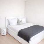 Schlafzimmer mit weißem Bett, grauer Decke und Nachttisch.