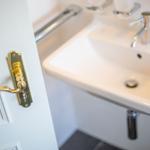 Hinter einer weißen Tür mit goldenem Griff ist ein Waschbecken mit Wasserhahn sichtbar.