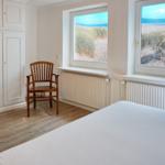 Schlafzimmer mit Blick auf Strand und Meer, weißes Bett, Holzstuhl und Kleiderschrank.
